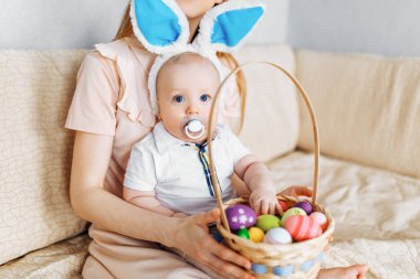 Mutlu Paskalyalar. Paskalya günü tavşan kulaklı bir anne ve küçük bir bebek, Paskalya yumurtası boyalı, eğlen, oyna ve sarıl, kanepede otur. Aile Paskalyayı kutluyor