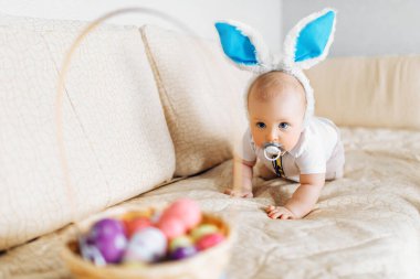 Tavşan kulaklı sevimli bebek evde kanepenin üzerinde paskalya yumurtalarıyla sepete doğru sürünüyor.