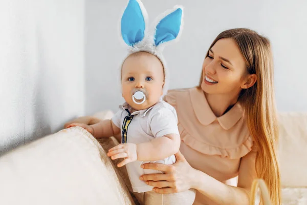 ママとウサギの耳を持つ赤ちゃん イースターエッグを手に ソファに座って 両親と子供たちは屋内でプレイします 家族はイースターを祝う — ストック写真
