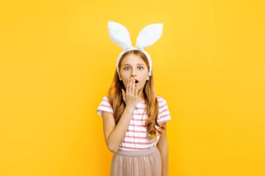 Şok olmuş, küçük güzel bir kızın kafasına tavşan kulağı takılmış, şaşırmış, ağzı sarı bir arka planda, eliyle kapatılmış. Paskalya ve baharın sembolü.