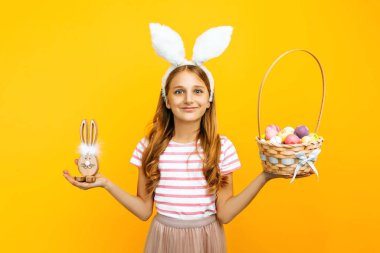 Tavşan kulaklı mutlu güzel bir kız elinde renkli yumurtalarla hasır bir sepet ve sarı arka planda tahta bir Paskalya Tavşanı tutuyor. Paskalya ve baharın sembolü