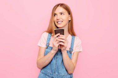 Gülümseyen, şık bir genç kadının portresi günlük giysiler içinde, elinde kahveyle, izole edilmiş pembe bir arka planda poz veriyor.