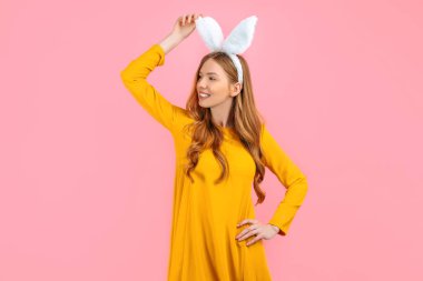 Holiday season of rabbit and eggs. A happy stylish woman in Easter Bunny ears isolated on a pink background looks at the space. clipart