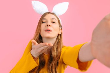 Mutlu Paskalyalar. Paskalya Tavşanı kulaklarıyla gülümseyen şık bir kadın telefonuyla selfie çekiyor ve izole edilmiş pembe bir arka planda öpüşüyor.