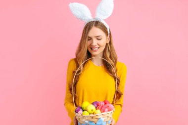 Happy Easter. A beautiful happy young woman wears rabbit ears on Easter day and holds a basket of Easter eggs, on an isolated pink background. Beautiful girl is getting ready for Easter clipart
