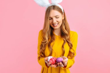 The concept of a happy Easter. A happy young girl with rabbit ears holds colorful Easter eggs, on a pink background. clipart