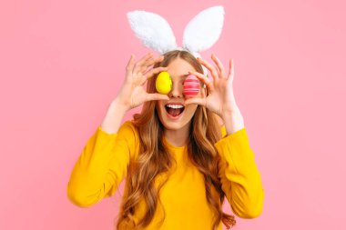 Tavşan kulağı takan ve gözünün önünde renkli Paskalya yumurtaları tutan mutlu genç bir kadın.