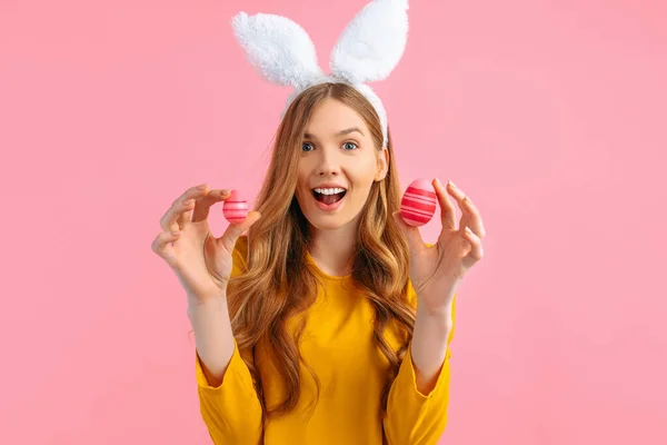 The concept of a happy Easter. Happy, surprised, shocked girl with rabbit ears holding colorful Easter eggs, on a pink background.