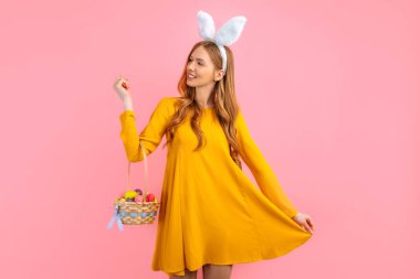 Happy Easter. A beautiful happy young woman wears rabbit ears on Easter day and holds a basket of Easter eggs, on an isolated pink background. Beautiful girl is getting ready for Easter clipart