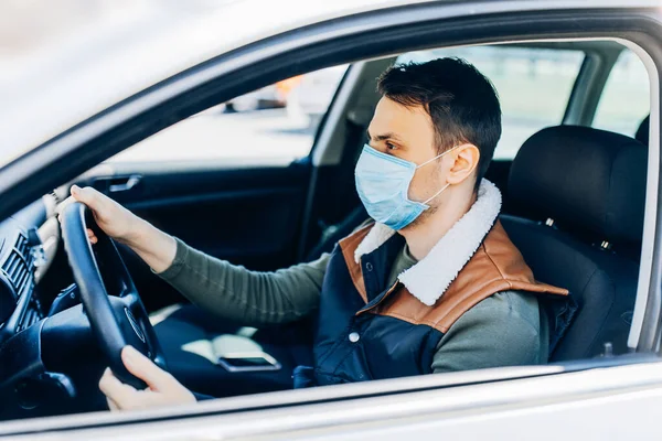 Beautiful young masked man sitting in a car, protective mask against coronavirus, driver on a city street during a coronavirus outbreak, covid-19