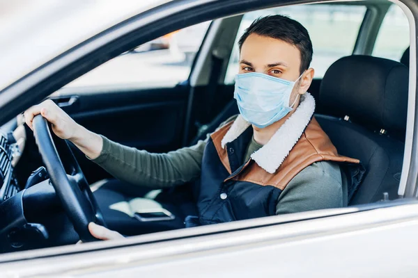 Beautiful young masked man sitting in a car, protective mask against coronavirus, driver on a city street during a coronavirus outbreak, covid-19