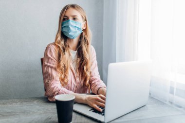 Genç bir iş kadını evde çalışıyor, bir dizüstü bilgisayarla masada oturuyor, koruyucu bir maske takıyor. Karantina, koronavirüs konsepti