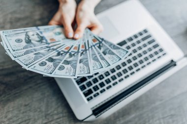 Bilgisayarın arka planında dolar banknotlarını tutan ellerin yakın çekimi. Uzaktan çalışma, serbest çalışma, online alışveriş kavramı