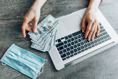Bilgisayarın arka planına karşılık dolar banknotları ve virüslerden korunan bir maske tutan ellerin yakın çekimi. Uzaktan çalışma kavramı, karantina, koronavirüs, online alışveriş
