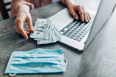 Bilgisayarın arka planına karşılık dolar banknotları ve virüslerden korunan bir maske tutan ellerin yakın çekimi. Uzaktan çalışma kavramı, karantina, koronavirüs, online alışveriş