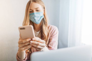 Koruyucu maskeli genç bir iş kadını karantina sırasında evinde çalışıyor ve cep telefonuyla masada oturuyor. Karantina, koronavirüs,