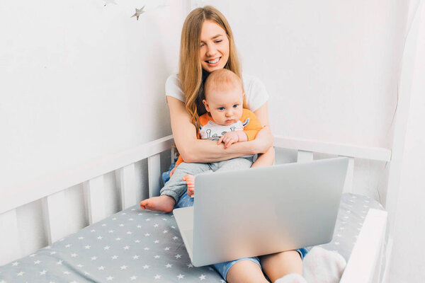 Молодая мать в пижаме с маленьким ребенком, отдыхающим на кровати, с портативным ноутбуком
