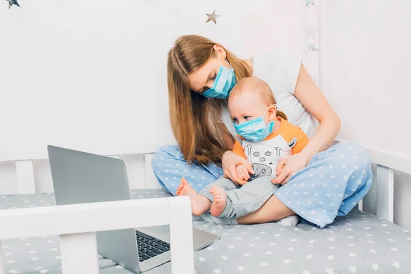 Evde pijama giyen ve yüzünde tıbbi koruyucu bir maske takan genç bir anne ve yatağında taşınabilir bir dizüstü bilgisayarla oturan küçük bir çocuk. Karantina ve koronavirüsteki uzaktan kumanda konsepti
