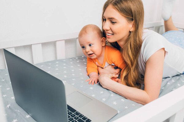 A young mother in pajamas with a small child resting on the bed, with a portable laptop