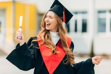 Yüksek lisans elbisesi içinde, elinde üniversiteden diploması olan mutlu, güzel bir kız öğrenci. Mezuniyet töreni konsepti