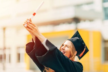 Yüksek lisans elbisesi içinde, elinde üniversiteden diploması olan mutlu, güzel bir kız öğrenci. Mezuniyet töreni konsepti