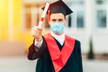Erkek bir lisansüstü öğrencisi koronavirüse karşı koruyucu bir maske takıyor, elinde diplomasıyla siyah bir mezuniyet elbisesi içinde. Mezuniyet töreni kavramı, karantina, koronavirüs.