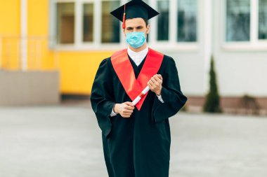 Erkek bir lisansüstü öğrencisi koronavirüse karşı koruyucu bir maske takıyor, elinde diplomasıyla siyah bir mezuniyet elbisesi içinde. Mezuniyet töreni kavramı, karantina, koronavirüs.