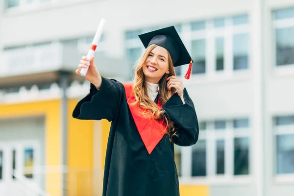 Yüksek lisans elbisesi içinde, elinde üniversiteden diploması olan mutlu, güzel bir kız öğrenci. Mezuniyet töreni konsepti