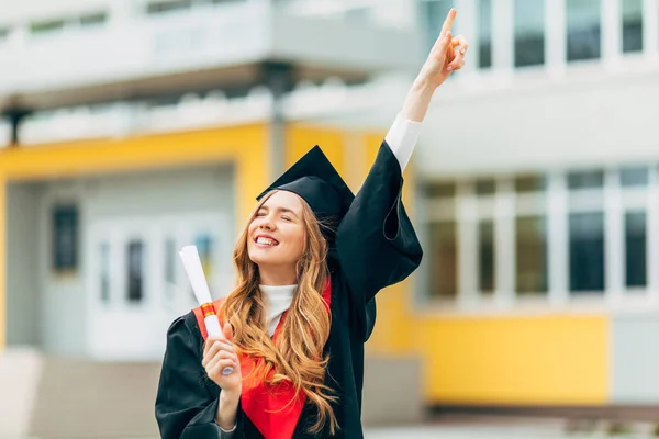 Yüksek lisans elbisesi içinde, elinde üniversiteden diploması olan mutlu, güzel bir kız öğrenci. Mezuniyet töreni konsepti
