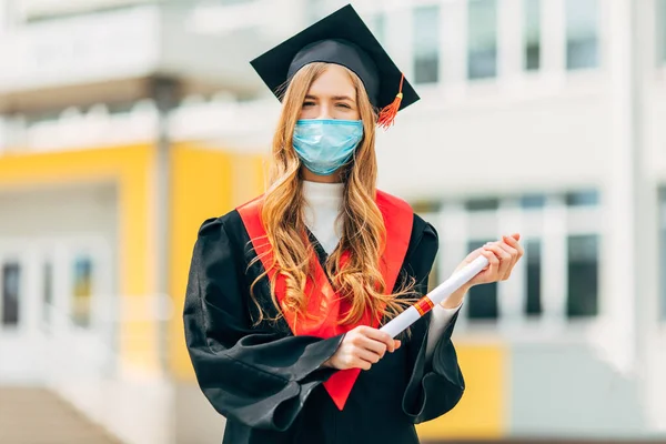 A female graduate student in a protective medical mask, in a black graduation dress, with a diploma in her hands. Graduation ceremony concept, quarantine, coronavirus