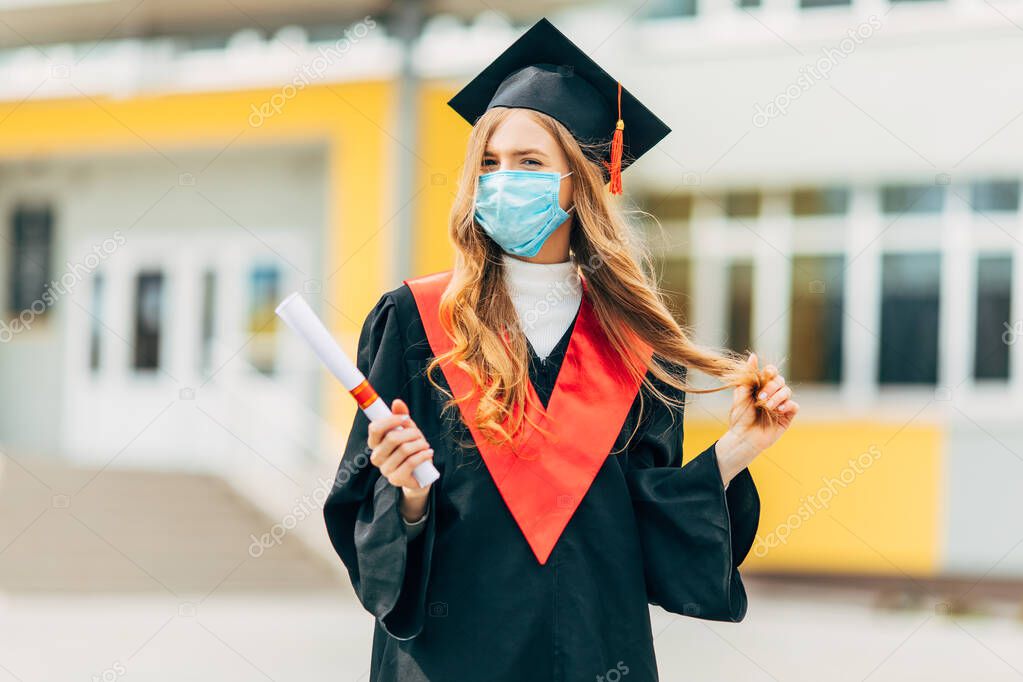 A female graduate student in a protective medical mask, in a black graduation dress, with a diploma in her hands. Graduation ceremony concept, quarantine, coronavirus