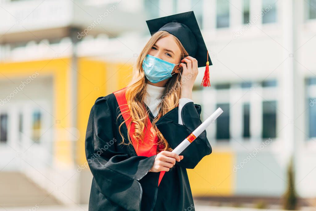 A female graduate student in a protective medical mask, in a black graduation dress, with a diploma in her hands. Graduation ceremony concept, quarantine, coronavirus