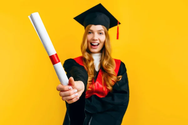 Happy attractive graduate in a master\'s dress, with a diploma on a yellow background. Concept of the graduation ceremony