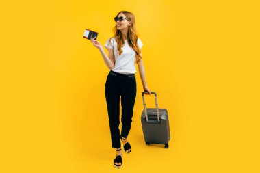 Happy beautiful woman, girl with a suitcase and passport with tickets on a yellow background. The concept of travel, business clipart