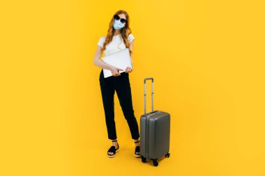 Siyah gözlüklü genç bir iş kadını yüzünde koruyucu bir tıbbi maske, bir dizüstü bilgisayar ve sarı arka planda bir bavul. Seyahat, yurt dışına seyahatler, karantina ve koronavirüs.