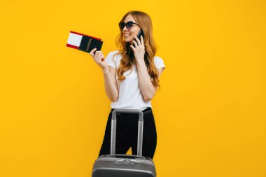 Siyah gözlüklü mutlu bir genç kadın, cep telefonuyla konuşuyor, elinde bavul ve sarı arka planda biletleri olan bir pasaportla duruyor. Seyahat, iş konsepti