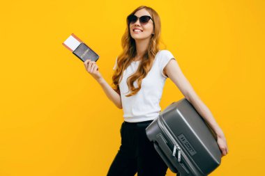 Güneş gözlüklü gülümseyen bir kadın yolcu, sarı arka planda izole edilmiş bir bavul ve pasaportla ayakta duruyor. Seyahat kavramı