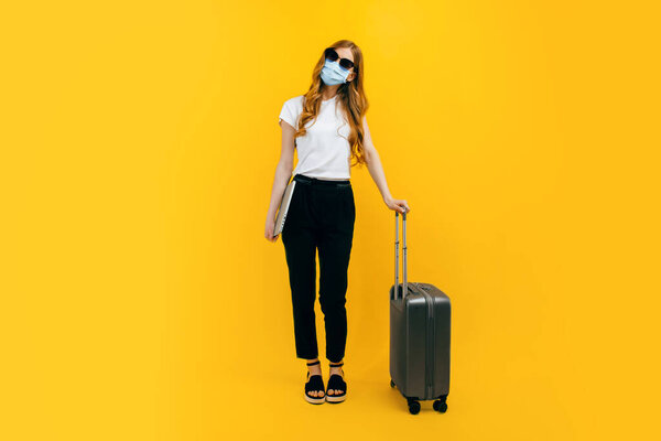 Молодая деловая женщина в темных очках, защитная медицинская маска на лице, ноутбук и чемодан на желтом фоне. Концепция путешествий, поездок за границу, карантина и коронавируса
