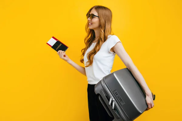 一个戴着太阳镜 面带微笑的女乘客站在那里 手里拿着手提箱和护照 车票被黄色背景隔开 旅行概念 — 图库照片