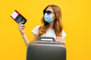 Yüzünde tıbbi koruyucu maske takan, elinde biletler ve pasaportla sarı arka planda duran bir kadın yolcu. Karantina, koronavirüs, seyahat kavramı