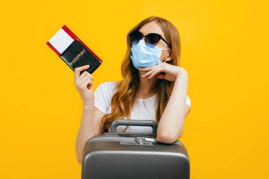 Yüzünde tıbbi koruyucu maske takan, elinde biletler ve pasaportla sarı arka planda duran bir kadın yolcu. Karantina, koronavirüs, seyahat kavramı