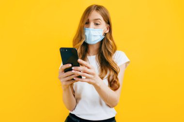 Koruyucu tıbbi maske takan genç bir kadın sarı arka planda cep telefonu kullanıyor. Karantina, koronavirüs konsepti