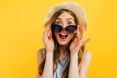 Şok olmuş güneş gözlüklü ve sarı arka planda yaz şapkalı bir kız. Seyahat, tatil, yaz