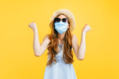 Yazın şapkalı bir kızın portresi, viral enfeksiyona karşı tıbbi koruyucu bir maske takıyor, sarı izole bir arka planda zaferi kutluyor. Karantina, koronavirüs, yaz