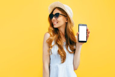 Yazlık şapkalı mutlu güzel kız mavi arka planda fotokopi alanı için boş bir cep telefonu gösteriyor.