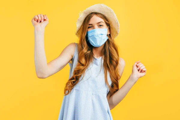 Portrait of a girl in a hat in summer, wears a medical protective mask against a viral infection, celebrates victory on a yellow isolated background. Quarantine, coronavirus, summer