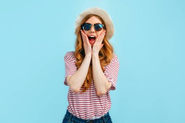 Güneş gözlüklü şaşırmış kız. Gözlükler ve yaz şapkası, kız ellerini izole edilmiş mavi bir arka planda tutarak ağzını kapatıyor.