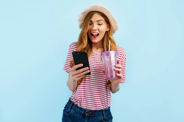 戴着夏帽 站在那里喝着鸡尾酒 上网或使用手机 背景是蓝色的快乐女孩 — 图库照片