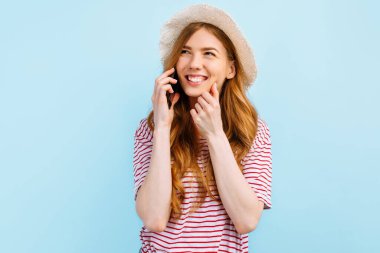 Yazlık şapkalı, cep telefonuyla konuşan, mavi arka planda duran düşünceli kız.
