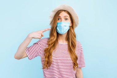 Yazlık şapkalı bir kız mavi arka planda yüzüne koruyucu maske takıyor. Karantina, koronavirüs, yaz tatili konsepti